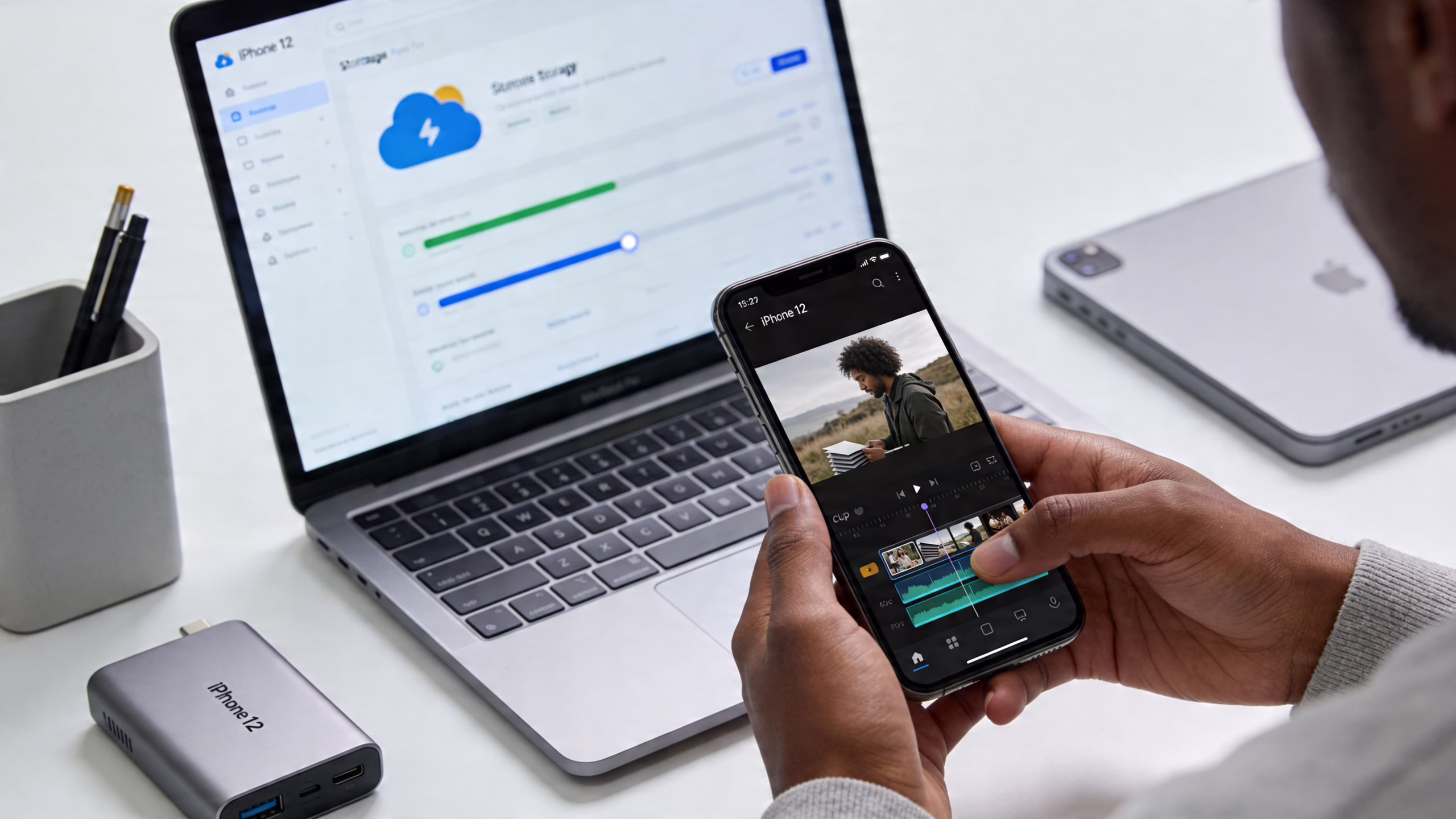 A person uses their iPhone 12 to edit a video while a laptop displays storage settings nearby.